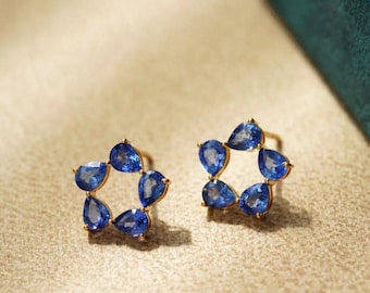 May include: A pair of gold stud earrings, each featuring a starburst design of five pear-shaped, vibrant blue gemstones. The earrings are set against a neutral background, highlighting the jewelry's color and design.