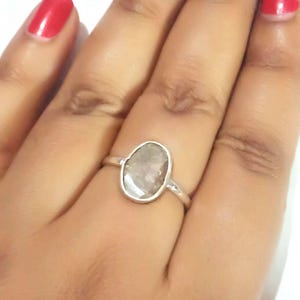 May include: A silver ring featuring an oval, pale gemstone. The ring has a simple, polished silver band. The gemstone is approximately 1.9 cm long and 1.3 cm wide. The ring is displayed on a hand with red nail polish.