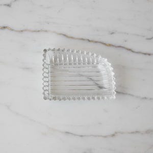 Puede incluir: Un plato de vidrio transparente, rectangular, con un borde decorativo festoneado. El plato tiene un interior acanalado y se asienta sobre una superficie de mármol blanco con vetas doradas. El plato probablemente se utiliza para servir o exhibir.