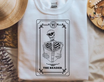 Può includere: Felpa bianca con un disegno di carta dei tarocchi in bianco e nero. La carta raffigura uno scheletro con occhiali da sole che legge un libro, con la scritta "THE READER". Il disegno è incorniciato da bordi decorativi.