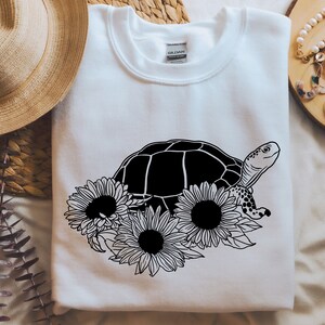 May include: A white sweatshirt with a black and white graphic of a turtle surrounded by sunflowers.