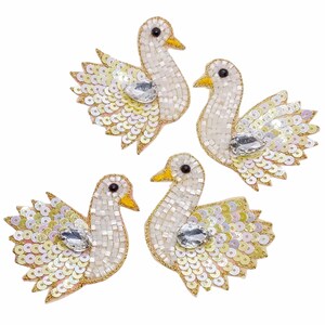 May include: Four decorative swan embellishments. Each swan is white with a yellow beak and black eyes. The wings are made of iridescent sequins, and a large, clear gem is on the chest. The swans are outlined in gold.