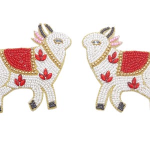 May include: Two beaded cow figurines. The cows are white with red and gold accents. They have red leaf-shaped embellishments and a red saddle. The horns and hooves are black. The background is white.