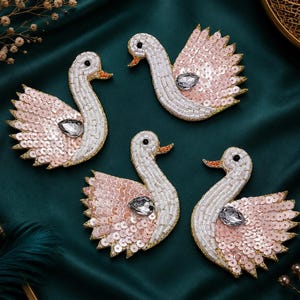 May include: Four swan-shaped patches featuring white beaded necks and heads with orange beaks and black eyes. The wings are crafted from light pink sequins with a single clear teardrop-shaped crystal accent in the centre of each wing.