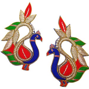 Peut inclure: Deux appliqués brodés de paon. Chaque appliqué présente un paon bleu avec une poitrine rouge, une queue rouge et verte, et des accents dorés et argentés. Les appliqués sont conçus avec des détails complexes et sont posés sur un fond blanc.