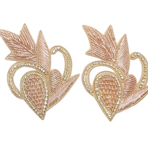 May include: Two ornate, symmetrical appliques with a floral design. The appliques feature a light peach-colored embroidered pattern, accented with clear rhinestones and a gold-colored border. The design includes leaf-like shapes and a heart-shaped element.