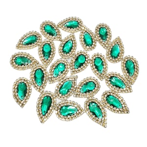 Könnte beinhalten: Eine Sammlung von smaragdgrünen, tropfenförmigen Strasssteinen mit goldfarbenem Rand und einer Umrandung aus kleinen, klaren Strasssteinen. Die Verzierungen sind auf weißem Hintergrund verstreut und zeigen ihre funkelnden Details.
