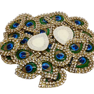Könnte beinhalten: Ein Haufen von Pfauenfeder-förmigen Verzierungen mit Strasssteinen und einem blauen und grünen Design. Die Verzierungen sind goldfarben und haben eine glänzende Oberfläche.