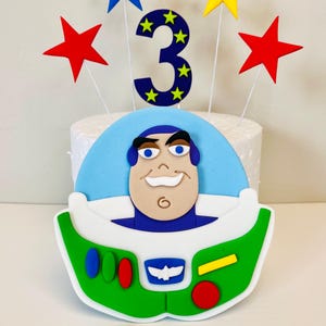 May include: A cake topper featuring Buzz Lightyear from Toy Story. The topper is made of fondant and includes a blue and yellow number 3, red and yellow stars, and a green and white spaceship. The topper is on a white cake with a blue fondant base. There are also six white fondant clouds on a blue background.