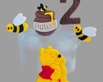 Pode incluir: Conjunto de topo de bolo com tema do Winnie the Pooh com uma figura amarela do Winnie the Pooh, um pote de mel marrom com uma abelha no topo, um número 2 marrom e duas abelhas amarelas e pretas.