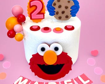 May include: A white cake decorated with Sesame Street characters, including Cookie Monster and Elmo. The cake is topped with a number 2 and the name "MABEL" in pink letters. Pink and orange decorative elements surround the cake.