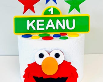 May include: A colorful cake topper featuring a red Elmo face, a green sign with the name "Keanu", and three star-shaped decorations. The sign also displays the number "1". Additional colorful circles are placed on the cake.