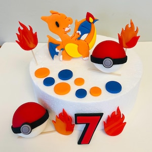May include: A white cake decorated with a red and white Pokeball, an orange and yellow Charizard Pokemon, orange and red flames, and a red number 7. The cake is also decorated with orange and blue circles.