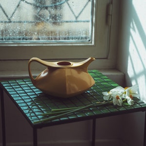 Puede incluir: Una tetera de cerámica amarilla se encuentra sobre una mesa con azulejos verdes y un ramo de narcisos blancos.