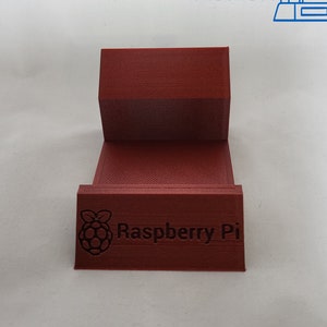 May include: A red 3D printed stand with the text "Raspberry Pi" on the front. The stand is designed to hold a Raspberry Pi computer.