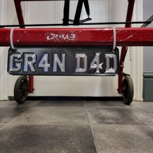 May include: A black number plate with silver lettering that reads "GR4N D4D". The plate is hanging from a red metal bar.