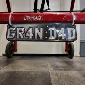 May include: A black number plate with silver lettering that reads "GR4N D4D" with iridescent stars scattered across the plate.
