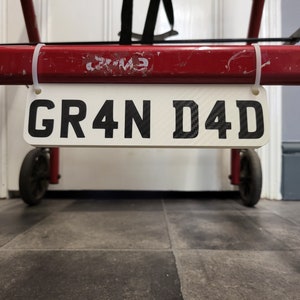 May include: A rectangular white sign displaying the text "GR4N D4D" in black, attached to a red metal frame. The sign is secured with white cable ties. The background features a tiled floor and a white door.