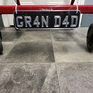 May include: A black license plate with the text "GR4N D4D" in silver lettering. The plate is attached to a red metal frame with white cable ties. Small colourful star-shaped accents are visible.
