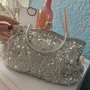May include: A silver clutch bag featuring intricate beadwork and sequin embellishments in a floral pattern. The bag has a metallic frame, a kiss-lock closure, and a rounded silver handle. The surface is covered in shimmering silver beads and sequins.