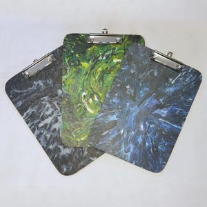 May include: Three clipboards with different abstract designs. The clipboards are made of wood and have a silver metal clip. The designs are a swirling green and yellow, a swirling blue and black, and a swirling black and white.