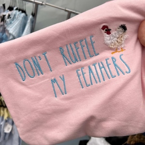 May include: A light pink long-sleeved shirt with the embroidered phrase "DON'T RUFFLE MY FEATHERS" in light blue thread. A small, colorful embroidered chicken is on the sleeve. The shirt is displayed in a retail setting.