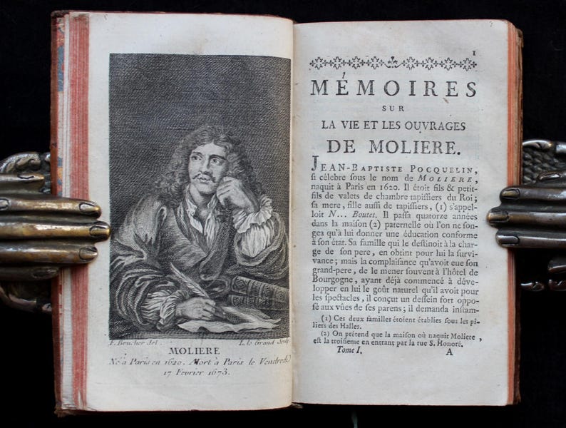 K&ouml;nnte beinhalten: Ein aufgeschlagenes antikes Buch mit einer detaillierten Gravur von Moli&egrave;re auf der linken Seite. Die rechte Seite zeigt franz&ouml;sischen Text. Das Buch wird von einem dekorativen, handf&ouml;rmigen Buchhalter aus Metall offen gehalten.