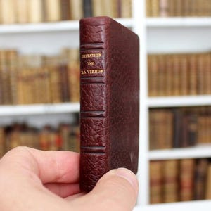 Puede incluir: Un libro pequeño encuadernado en cuero color burdeos oscuro con una superficie texturizada. El lomo presenta bandas horizontales en relieve y texto dorado en mayúsculas que dice "IMITATION DE LA VIERGE" en orientación vertical.
