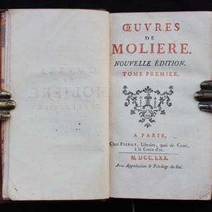 K&ouml;nnte beinhalten: Ein offenes Buch mit dem Titel "&OElig;UVRES DE MOLIERE" in roter und schwarzer Tinte. Das Buch wird von einem dekorativen Buchhalter aus Metall offen gehalten. Der Text enth&auml;lt "NOUVELLE &Eacute;DITION" und "TOME PREMIER."
