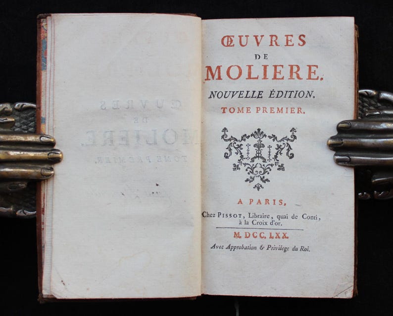 K&ouml;nnte beinhalten: Ein offenes Buch mit dem Titel "&OElig;UVRES DE MOLIERE" in roter und schwarzer Tinte. Das Buch wird von einem dekorativen Buchhalter aus Metall offen gehalten. Der Text enth&auml;lt "NOUVELLE &Eacute;DITION" und "TOME PREMIER."