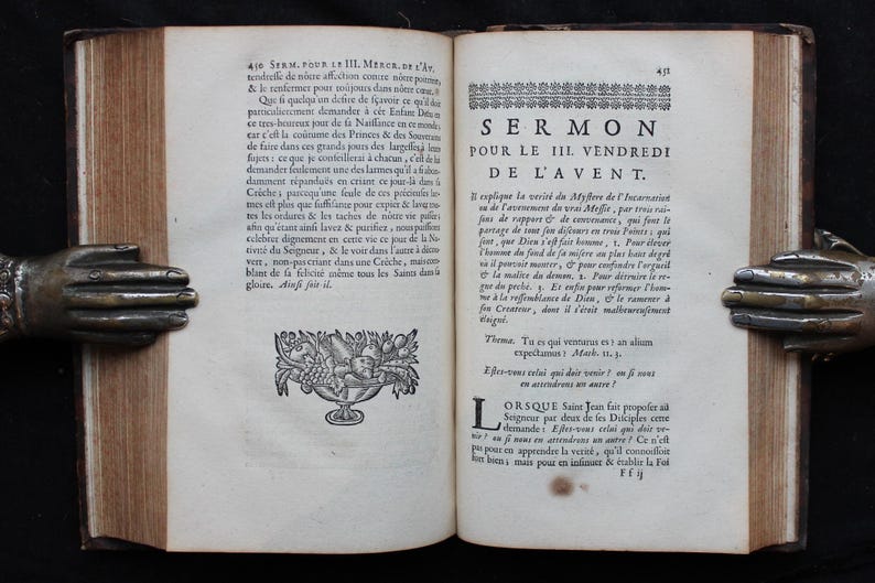 May include: An open antique book with text and illustrations, held by bronze hands. The pages display French text, with the title "SERMON POUR LE III VENDREDI DE L'AVENT."