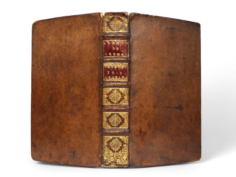 May include: Antique book with a brown leather cover. The spine is decorated with gold filigree and red panels with the text "SERMON DE GRENAD" and "T.O.I". The book is aged and shows signs of wear.