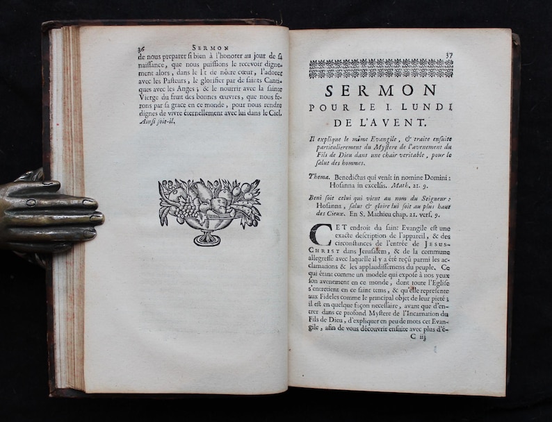 May include: An open book with text and illustrations. The left page has a decorative bowl of fruit. The right page features the title "SERMON POUR LE I LUNDI DE L'AVENT" in large text. The book is held by a bronze hand.