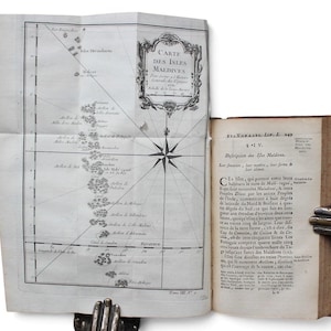 May include: An antique book open to a page with a fold-out map titled Carte des Isles Maldives. The map features a compass rose and detailed geographical markings. The adjacent page contains printed French text with the header Description des Isles Maldives.