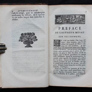 May include: Open book with aged pages, featuring French text and decorative illustrations. The left page has a floral design in a vase. The right page has a title and text. The book is held by a bronze hand.