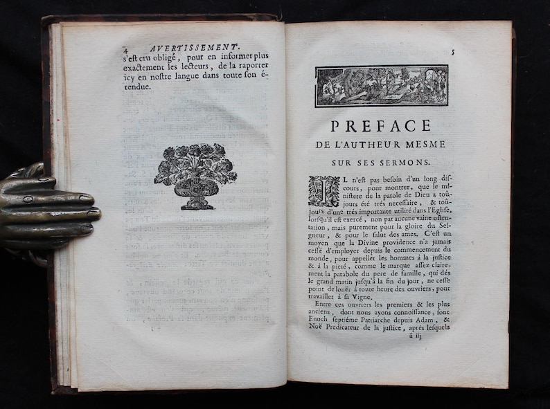 May include: Open book with aged pages, featuring French text and decorative illustrations. The left page has a floral design in a vase. The right page has a title and text. The book is held by a bronze hand.