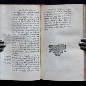 Pode incluir: Um livro antigo aberto com uma capa de couro marrom e p&aacute;ginas de cor creme. O livro est&aacute; aberto em uma p&aacute;gina com texto em franc&ecirc;s. O texto trata sobre a f&eacute; crist&atilde; e a import&acirc;ncia de ser fiel a Deus. O livro &eacute; mantido aberto por duas m&atilde;os met&aacute;licas decorativas.