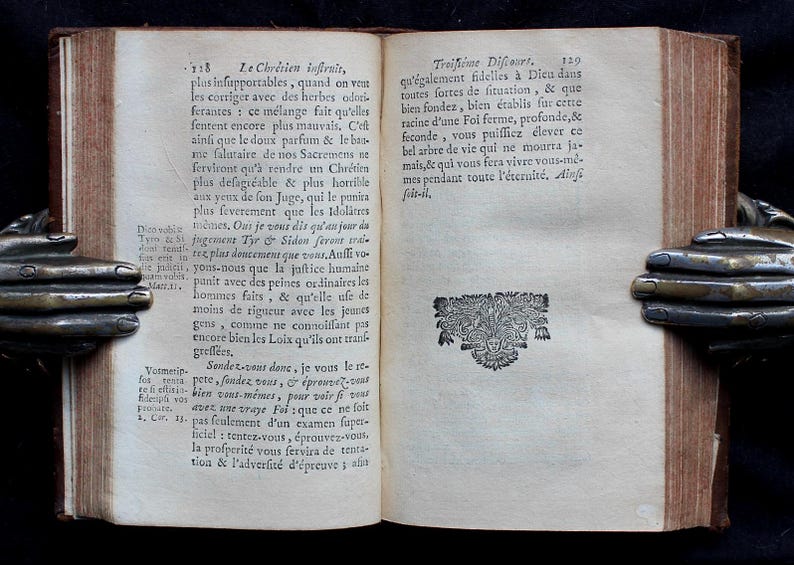Pode incluir: Um livro antigo aberto com uma capa de couro marrom e p&aacute;ginas de cor creme. O livro est&aacute; aberto em uma p&aacute;gina com texto em franc&ecirc;s. O texto trata sobre a f&eacute; crist&atilde; e a import&acirc;ncia de ser fiel a Deus. O livro &eacute; mantido aberto por duas m&atilde;os met&aacute;licas decorativas.