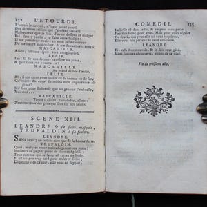 K&ouml;nnte beinhalten: Offenes antikes Buch mit franz&ouml;sischem Text, gehalten von einem Metallbuchhalter. Die Seiten sind gealtert und der Text in einer klassischen Schrift. Dekorative Elemente sind sichtbar. Das Buch scheint ein Theaterst&uuml;ck zu sein.