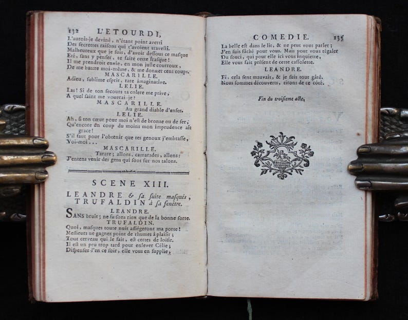 K&ouml;nnte beinhalten: Offenes antikes Buch mit franz&ouml;sischem Text, gehalten von einem Metallbuchhalter. Die Seiten sind gealtert und der Text in einer klassischen Schrift. Dekorative Elemente sind sichtbar. Das Buch scheint ein Theaterst&uuml;ck zu sein.