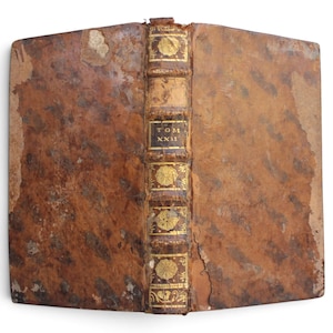 Op de afbeelding: Een antiek boek met een verweerde, gevlekte bruine leren kaft. De rug is voorzien van ribben, gouden bloemmotieven en een zwart label met de goudkleurige tekst "TOM XXII". De randen van de kaft vertonen duidelijke tekenen van slijtage.