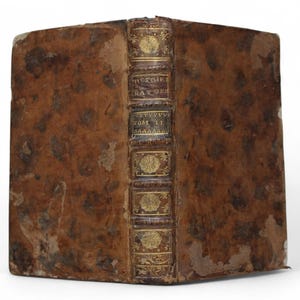 Pode incluir: Livro antigo com capa de couro castanho desgastada. A lombada apresenta letras douradas e elementos decorativos, com o título "HISTOIRE NATURELLE" e "TOM II" visíveis. As bordas do livro mostram sinais de idade e desgaste.
