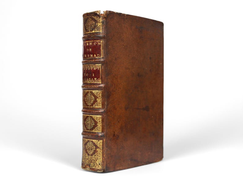 May include: A vintage, leather-bound book with gold accents. The spine features red panels with gold lettering and decorative elements. The book's cover is a rich brown color, and the edges show signs of wear, suggesting its age.
