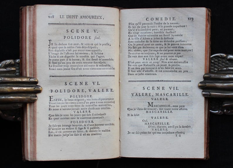 K&ouml;nnte beinhalten: Ein offenes antikes Buch mit gealterten, vergilbten Seiten. Der Text ist auf Franz&ouml;sisch, mit dem Titel "LE DEPIT AMOUREUX" sichtbar. Das Buch wird von verzierten Metallbuchst&uuml;tzen gehalten.