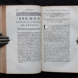 May include: An open antique book with text in French, held by bronze hands. The book's pages are aged and worn, with visible text and a decorative border. The book is a sermon.