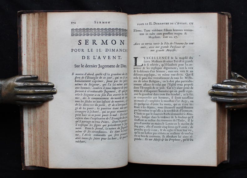 May include: An open antique book with text in French, held by bronze hands. The book's pages are aged and worn, with visible text and a decorative border. The book is a sermon.