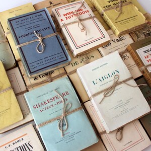 Puede incluir: Una colecci&oacute;n de libros antiguos, algunos con t&iacute;tulos franceses, est&aacute;n atados con cuerda y apilados sobre una superficie blanca. Los libros tienen varios colores, incluyendo amarillo, azul, blanco y marr&oacute;n.