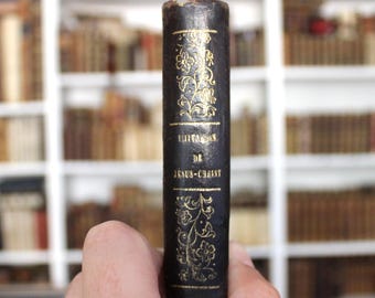 May include: A dark brown book spine featuring gold-colored floral patterns and the text "IMITATION DE JÉSUS-CHRIST" printed in gold lettering. The spine shows signs of wear at the top edge, with a vertical orientation and decorative filigree.
