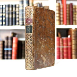 K&ouml;nnte beinhalten: Ein altes Buch mit einem abgenutzten, braunen Ledereinband. Der Buchr&uuml;cken hat goldene geometrische Muster und Text in Rot, Schwarz und Gold. Das Buch steht aufrecht, mit anderen alten B&uuml;chern auf Regalen im Hintergrund.