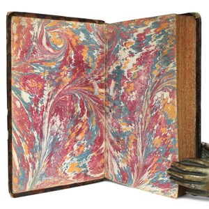 May include: An open antique book with marbled pages in shades of red, blue, yellow, and white. The book's cover is dark brown, and the pages are held open by a decorative metal hand. The book's pages have a swirling pattern.